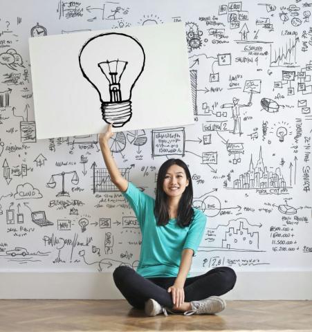 picture of a woman sitting on floor holding a b/w sign of a lightbulb above her head in front of a wall of B/W graphs