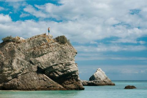picture of a person on a huge rock above the ocean with blue skies