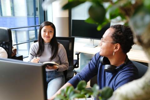 picture of two people looking at a computer monitor with one person taking notes in a paper notebook