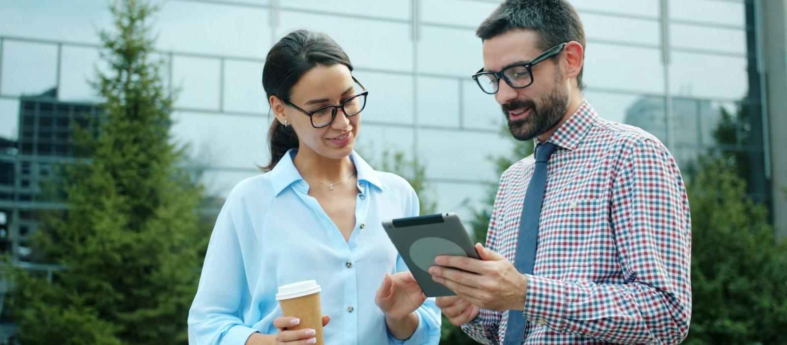 picture of two people with glasses and in professional clothes standing looking at a tablet 