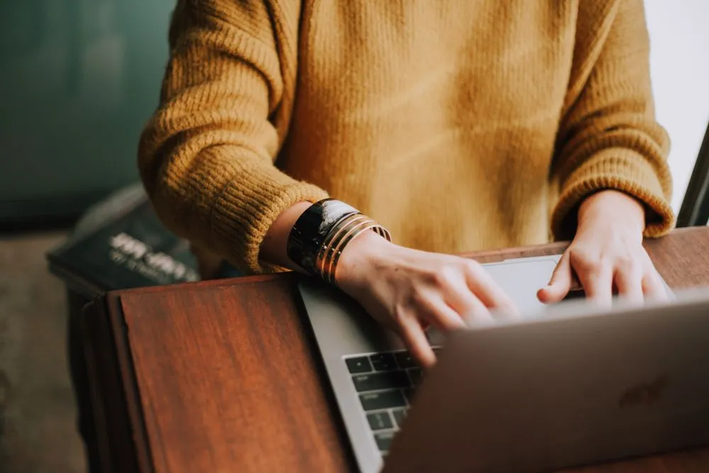 picture of person in a mustard sweater typing on a laptop computer with a window in the background