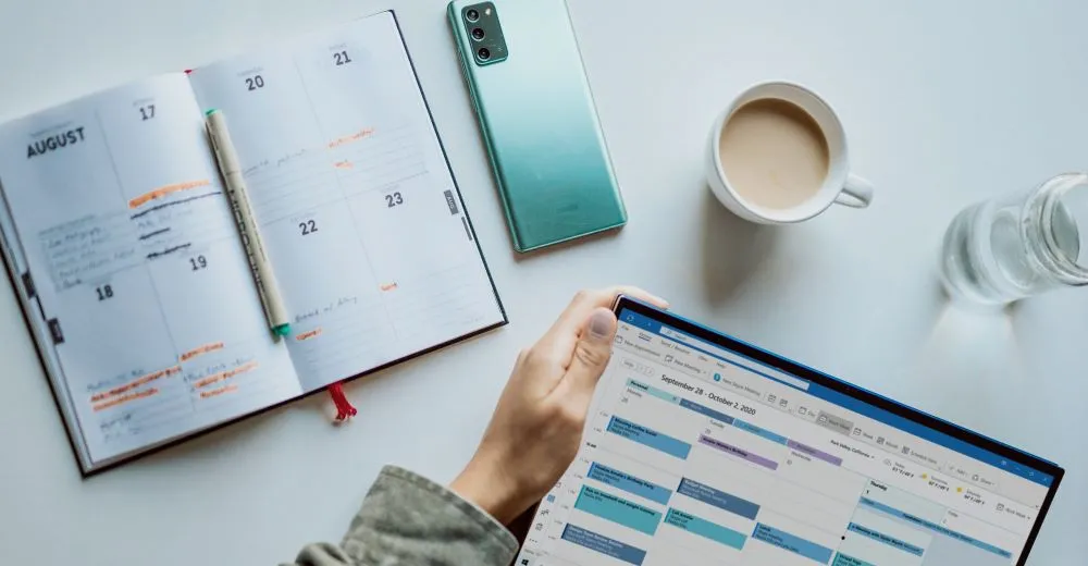 picture of desk with paper calendar opened to August and a computer with a calendar pulled up in schedule view